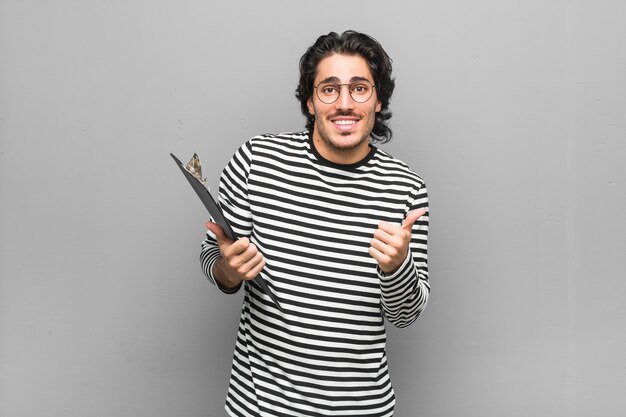 Hombre joven empleado sosteniendo un inventario levantando ambos pulgares, sonriente y confiado.