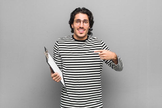 Hombre joven empleado sosteniendo un inventario sorprendido señalando con el dedo, sonriendo ampliamente.