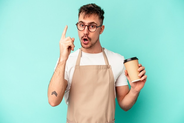 Hombre joven empleado de tienda sosteniendo un café aislado sobre fondo azul con una idea, concepto de inspiración.