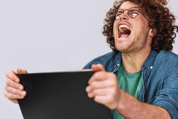 Hombre joven enojado con cabello rizado sentado en su escritorio con una computadora portátil que se siente estresado Hombre de negocios caucásico gritando mientras trabaja en la oficina con anteojos de moda posando sobre una pared blanca