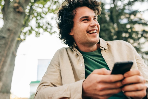 Hombre joven escribiendo mensajes en el teléfono móvil Hombre atractivo con cabello rizado sentado en el parque usando teléfono móvil y navegando en su teléfono inteligente