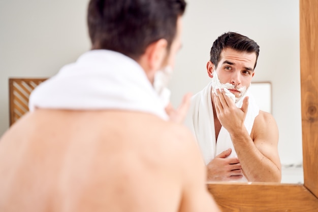 Hombre joven con espuma en la barba cerca del espejo en la mañana