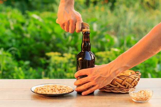 El hombre joven está abriendo una botella de cerveza con un abridor de edad. Botella de cerveza marrón con papas fritas en canasta de mimbre, cacahuetes en plato y recipiente sobre mesa de madera