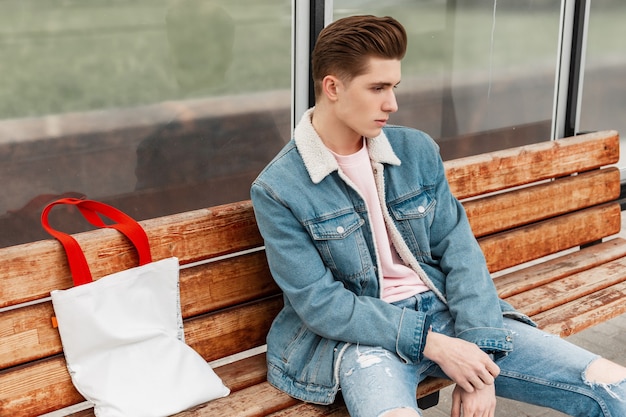 Hombre joven con estilo agradable en ropa de jeans casual juvenil azul de moda sentado en un banco de madera y esperando el transporte público. Chico guapo con ropa elegante en la parada de autobús de cristal. Moda masculina de verano.