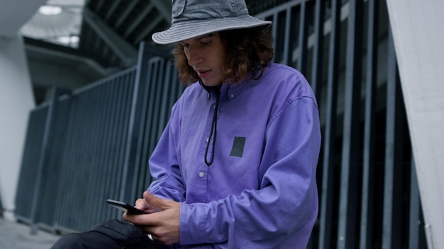 Hombre joven con estilo escribiendo en línea por teléfono móvil en el estadio de la ciudad Chico enfocado usando teléfono celular al aire libre Hipster guapo sentado en patineta con teléfono inteligente en las manos afuera