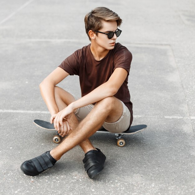 Hombre joven con estilo en gafas de sol sentado en una patineta.