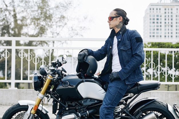 Hombre joven con estilo guapo en ropa de mezclilla sentado en motocicleta con casco en manos