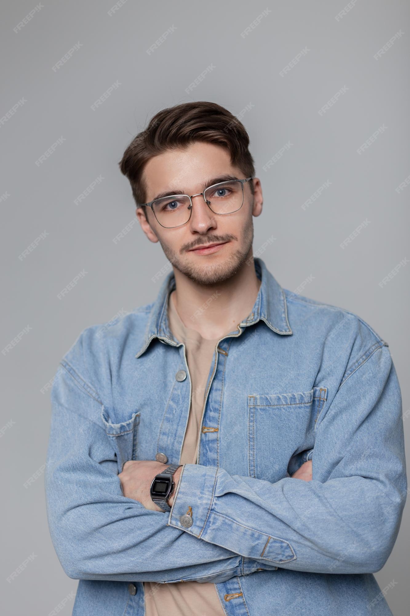Hombre joven con estilo hipster con gafas retro cabello en una camisa de mezclilla de moda sobre un fondo gris retrato de estudio de un chico guapo vintage Foto Premium