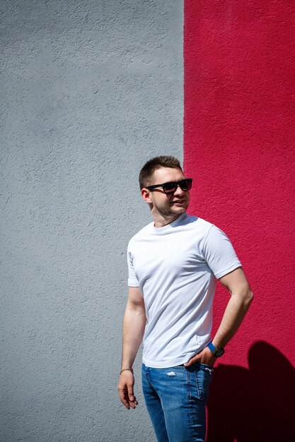 Hombre joven con estilo, un hombre vestido con una camiseta blanca en blanco de pie sobre un fondo de pared gris y rojo. Estilo urbano de ropa, imagen de moda moderna. Moda de hombres