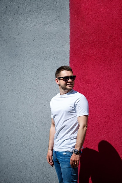 Hombre joven con estilo, un hombre vestido con una camiseta blanca en blanco de pie sobre un fondo de pared gris y rojo. Estilo urbano de ropa, imagen de moda moderna. Moda de hombres