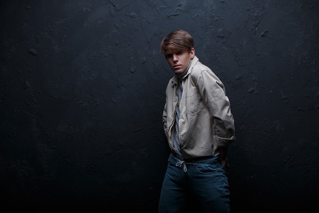 Hombre joven con estilo con un peinado de moda en una elegante chaqueta blanca con estilo en jeans con estilo vintage en golf gris se encuentra en un estudio oscuro cerca de una pared gris. Chico hermoso modelo de moda