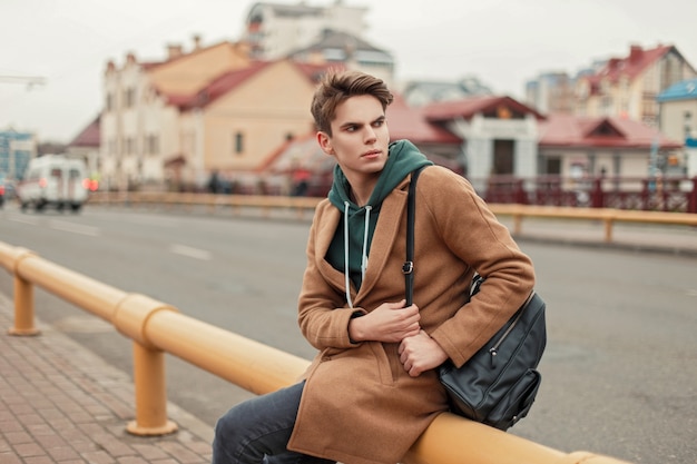 Hombre joven con estilo con el pelo en un abrigo de moda con capucha y bolso negro sentado en la calle en la ciudad