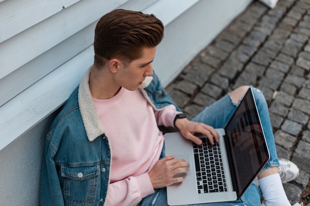 Hombre joven con estilo en sudadera rosa con ropa de jeans azul de moda trabaja de forma remota en la computadora portátil sentado en baldosas de piedra en la calle. Blogger exitoso trabaja, proyecto creativo. Tiempo de trabajo