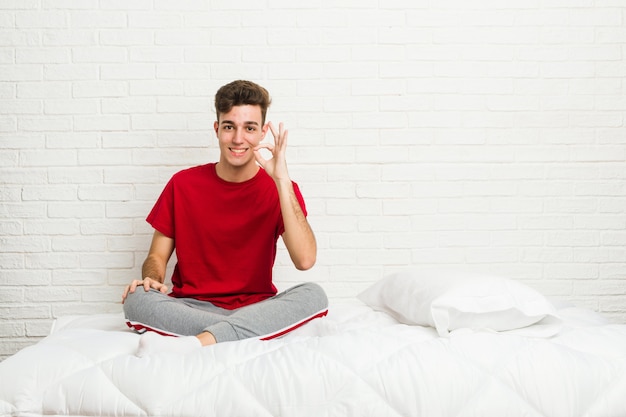 Hombre joven estudiante adolescente en la cama alegre y confiado mostrando gesto bien.