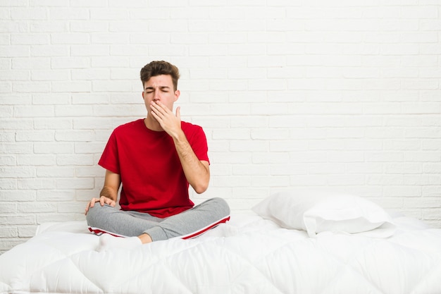 Hombre joven estudiante adolescente en la cama bostezando mostrando un gesto cansado cubriendo la boca con la mano.