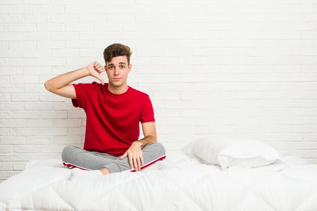 Hombre joven estudiante adolescente en la cama mostrando un gesto de disgusto, pulgares abajo. Concepto de desacuerdo