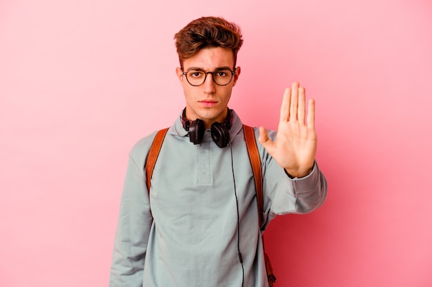 Hombre joven estudiante aislado sobre fondo rosa de pie con la mano extendida mostrando la señal de stop, impidiéndote.