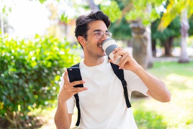 Hombre joven estudiante al aire libre mediante teléfono móvil y sosteniendo un café para llevar