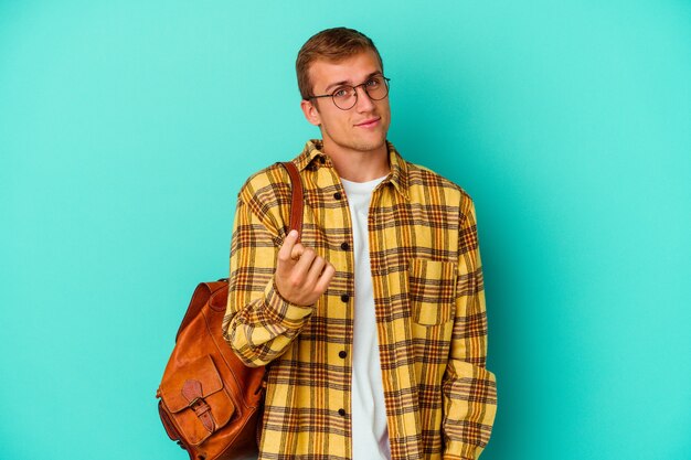 Hombre joven estudiante caucásico aislado en azul apuntando con el dedo como si invitara a acercarse.