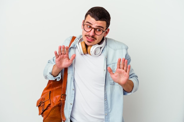 Hombre joven estudiante caucásico escuchando música aislado en la pared blanca rechazando a alguien mostrando un gesto de disgusto