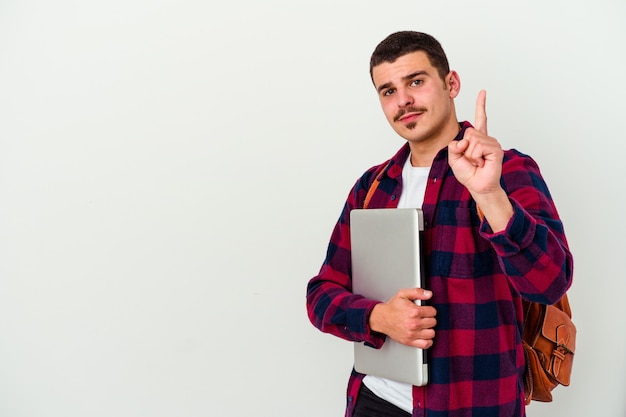 Hombre joven estudiante caucásico que sostiene una computadora portátil aislada en la pared blanca que muestra el número uno con el dedo.