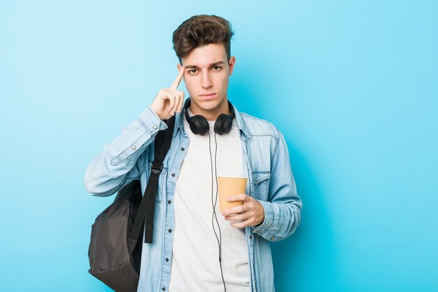 Hombre joven estudiante caucásico sosteniendo un café para llevar apuntando su sien con el dedo, pensando, centrado en una tarea.