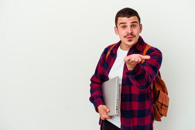 Foto hombre joven estudiante caucásico sosteniendo una computadora portátil aislada en la pared blanca doblando los labios y sosteniendo las palmas de las manos para enviar beso de aire.