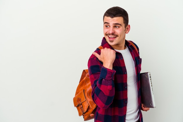 Hombre joven estudiante caucásico sosteniendo un portátil aislado sobre fondo blanco apunta con el dedo pulgar lejos, riendo y despreocupado.