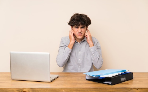 Hombre joven del estudiante con un ordenador portátil frustrado y que cubre los oídos