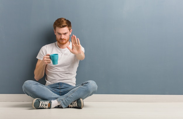 Hombre joven del estudiante del pelirrojo que se sienta en el piso que pone la mano en frente. Él está sosteniendo una taza de café.