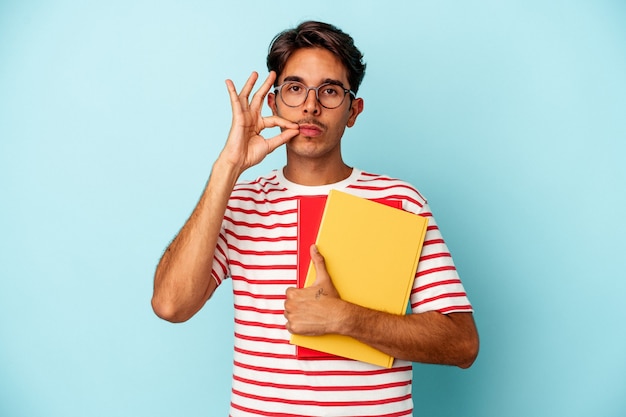 Hombre joven estudiante de raza mixta sosteniendo libros aislados sobre fondo azul con los dedos en los labios manteniendo un secreto.