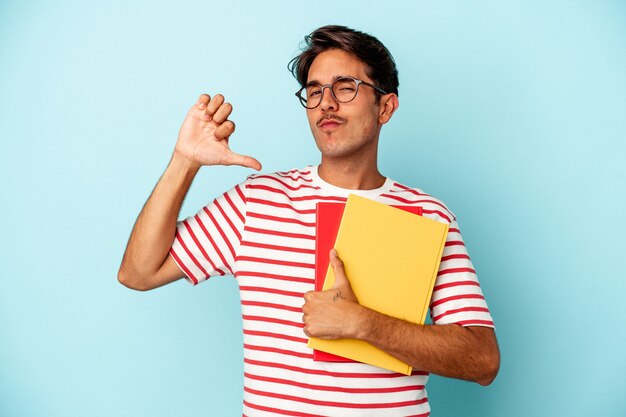 Hombre joven estudiante de raza mixta sosteniendo libros aislados sobre fondo azul se siente orgulloso y seguro de sí mismo, ejemplo a seguir.