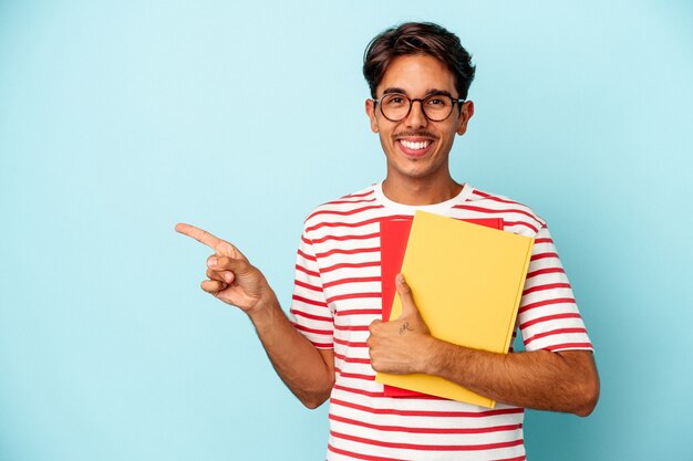 Hombre joven estudiante de raza mixta sosteniendo libros aislados sobre fondo azul sonriendo y apuntando a un lado, mostrando algo en el espacio en blanco.