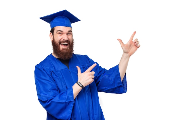 Hombre joven estudiante sorprendido en túnica azul graduándose y apuntando hacia otro lado