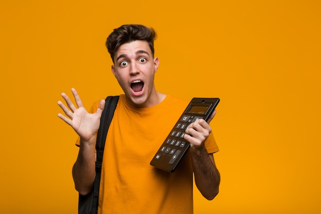 Hombre joven estudiante sosteniendo una calculadora feliz, sonriente y alegre.