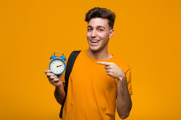Hombre joven estudiante sosteniendo un reloj despertador apuntando con el dedo hacia usted como invitando a acercarse.