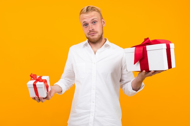 El hombre joven europeo rubio atractivo en una camisa blanca sostiene las cajas de regalo en un amarillo.