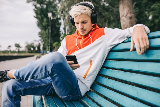 Hombre joven europeo rubio sincero disfrutando y escuchando música afuera en el parque Hombre guapo sentado en una silla y relajándose con música en auriculares bluetooth Concepto de personas de tecnología de estilo de vida