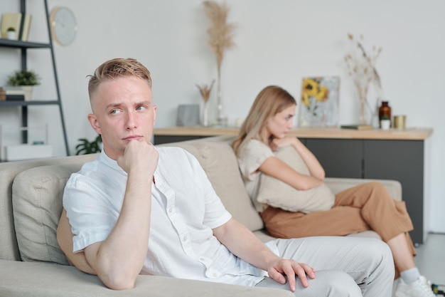 Hombre joven con expresión facial indiferente sentado en el sofá mientras ignora a su infeliz esposa o espera que ella hable primero durante la pelea
