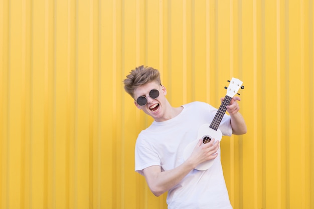 Hombre joven expresivo que juega el ukelele contra de una pared amarilla. Músico emocional toca el ukelele.