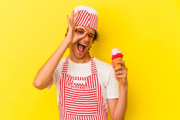 Hombre joven fabricante de helado con maquillaje sosteniendo helado aislado sobre fondo amarillo emocionado manteniendo el gesto ok en el ojo.