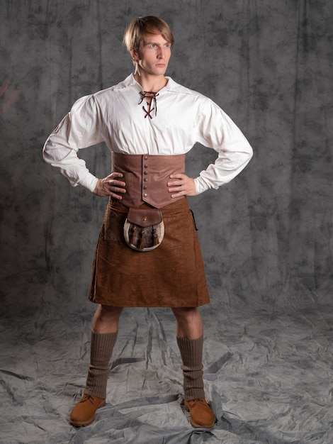 Un hombre joven con una falda escocesa de cuero y una blusa blanca con cordones. Un kilt escocés, marrón con un cinturón y sporran, una riñonera. Foto de cuerpo entero en el estudio sobre un fondo gris