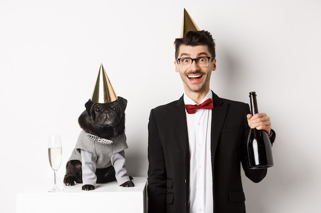 Foto hombre joven feliz celebrando con perro. lindo pug negro y hipster con conos de fiesta de cumpleaños, chico sosteniendo champán, de pie sobre fondo blanco.