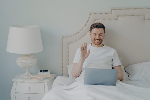 Hombre joven feliz con rastrojo agitando la mano a la computadora portátil mientras tiene una videollamada en línea y se sienta en la cama, conversando con sus amigos cercanos durante el tiempo libre el fin de semana en casa