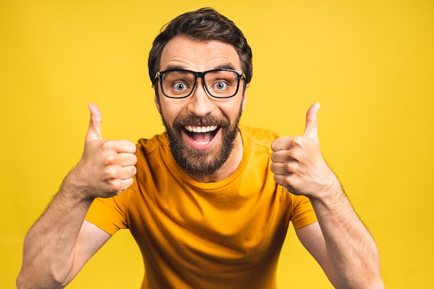 Hombre joven feliz. Retrato de hombre barbudo joven y guapo sonriendo mientras está de pie aislado sobre fondo amarillo. Pulgares hacia arriba.