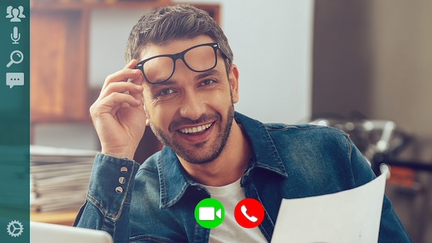 Hombre joven feliz en ropa casual elegante mirando a cámara y sonriendo mientras tiene videollamada