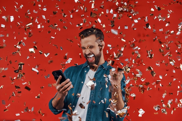 Hombre joven feliz en ropa casual sosteniendo un teléfono inteligente y gesticulando mientras está de pie