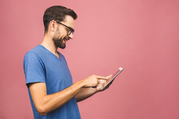 Hombre joven feliz en la situación casual y usar la tableta aislada sobre fondo rosado.