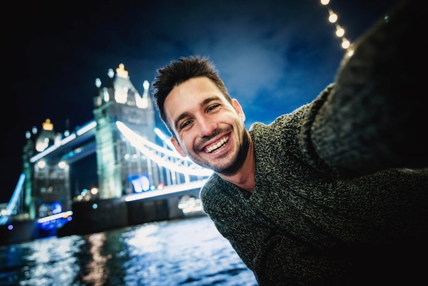 Hombre joven feliz tomando selfie en Tower Bridge Londres Chico guapo sonriendo a la cámara en las calles de la ciudad Turismo y concepto de estilo de vida feliz