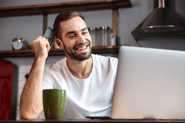 Hombre joven feliz usando la computadora portátil mientras está sentado en la mesa de la cocina, mostrando la tarjeta de crédito